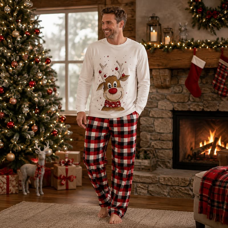 A man wearing a white long-sleeve pajama top with a reindeer graphic and red/black plaid pajama pants, standing barefoot in a cozy living room with a Christmas tree, fireplace, and stockings.